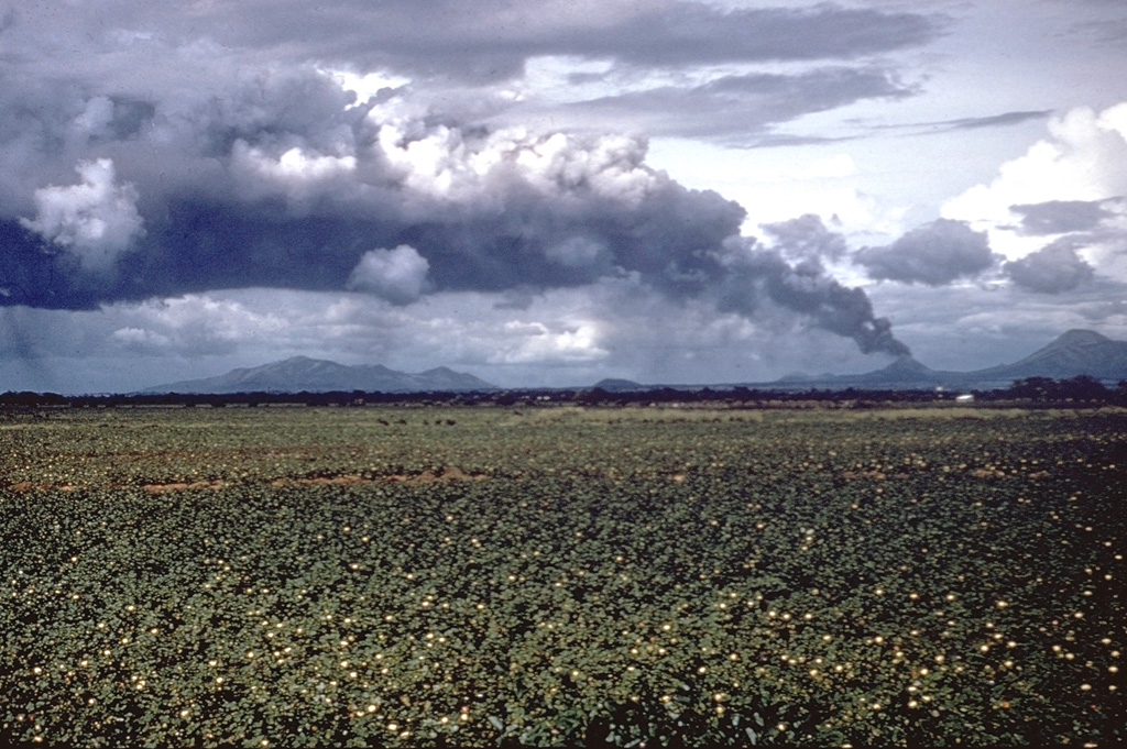 A large tephra cloud silhouetted against weather clouds rises from Cerro Negro volcano in November 1968.  Strong winds blowing from the east deflect the ash plume.  This view from the SW looks across cotton fields of the Nicaraguan central depression to the Marrabios Range volcanoes.  To the right of Cerro Negro is Las Pilas volcano, and the broad massif to the left is Rota volcano. Photo by Dick Stoiber, 1968 (Dartmouth College).