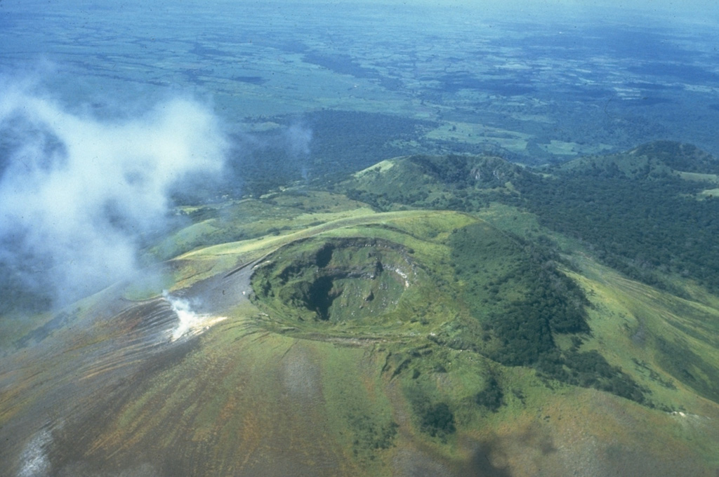 The summit crater of Las Pilas volcano (also known as El Hoyo) is 700 m wide and about 120 m deep.  A prominent fumarole on the SE side of the crater emits a steam plume.  Beyond and to the right of Las Pilas is the 900-m-high satellitic cone Cerro Ojo de Agua, itself capped with a 400-m-wide crater.  The diagonal line cutting across from the summit crater of Las Pilas to the lower right was formed during an eruption in 1952. Photo by Jaime Incer, 1981.