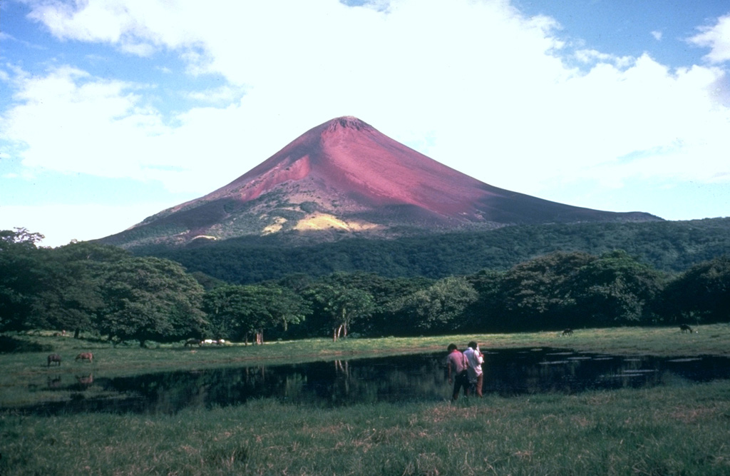 Oxidized, reddish-colored rocks form much of the summit cone of Momotombo volcano, seen here from the NW towering more than a kilometer above one of several small ponds on the floor of Monte Galán caldera.  The 4-km-wide caldera was the source of a major ignimbrite about 50,000 years ago. Photo by Jaime Incer.