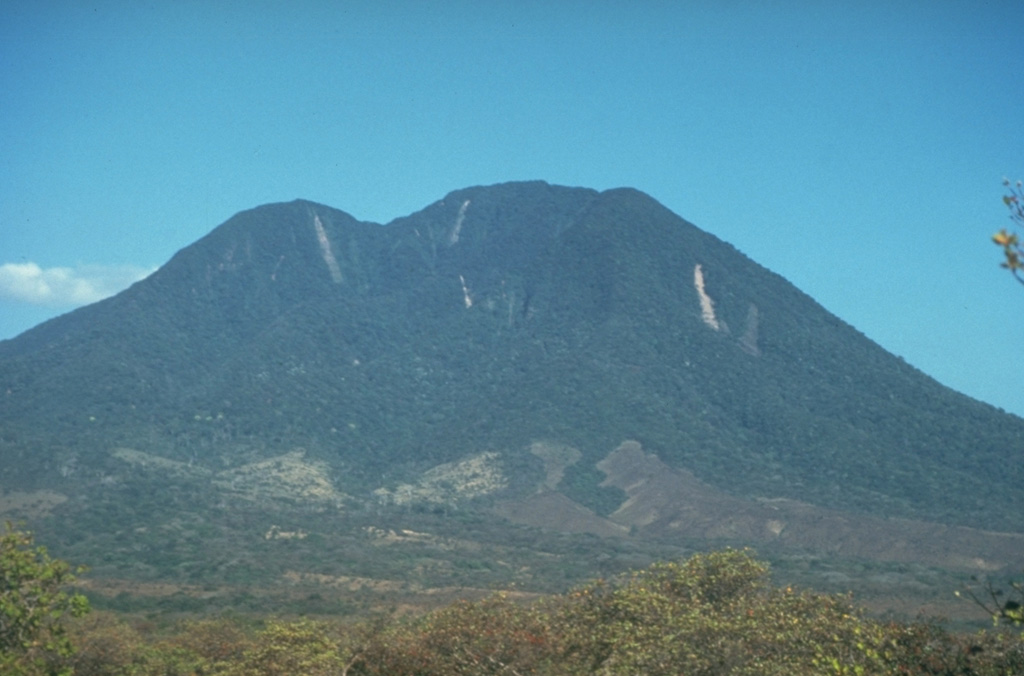 The SW side of Volcán Cacao, located at the SE end of the Orosí volcanic massif, is cut by an arcuate depression created during a massive slope failure in which the summit of the volcano was removed.  The vertical light-colored stripes on the upper edifice are rockslide scars.  The latest eruptive activity at the Orosí complex consisted of post-collapse lava domes and flows from Cerro Cacao. Photo by Cindy Stine, 1989 (U.S. Geological Survey).