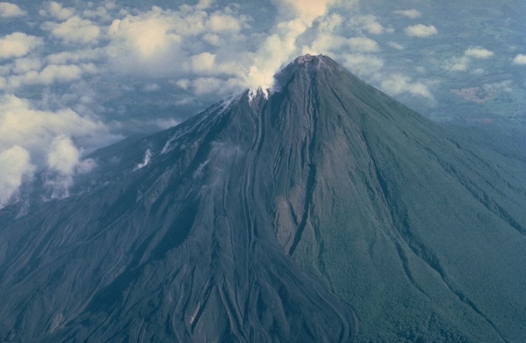 The symmetrical Volcán Arenal is the youngest stratovolcano in Costa Rica and one of its most active.  Steam rises from lava flows descending the west flank in this March 1983 view.  The oldest known products of Arenal volcano are only about 7000 years old.  Arenal's most recent eruptive period began with a major explosion in 1968.  Continuous explosive activity accompanied by slow lava effusion has occurred since from vents at the summit and western flank.     Copyrighted photo by Katia and Maurice Krafft, 1983.
