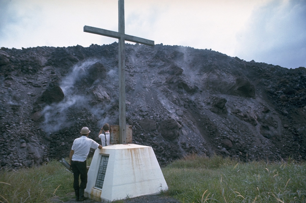 The margins of a lava flow descending the WNW flank of Arenal rise above a monument to victims of a powerful explosive eruption in July 1968. Lava flow effusion began on 19 September 1968 and by the time of this December 1968 photo had reached about 2 km from the vent on the lower west flank. Photo by Dick Berg, 1968 (courtesy of William Melson, Smithsonian Institution).