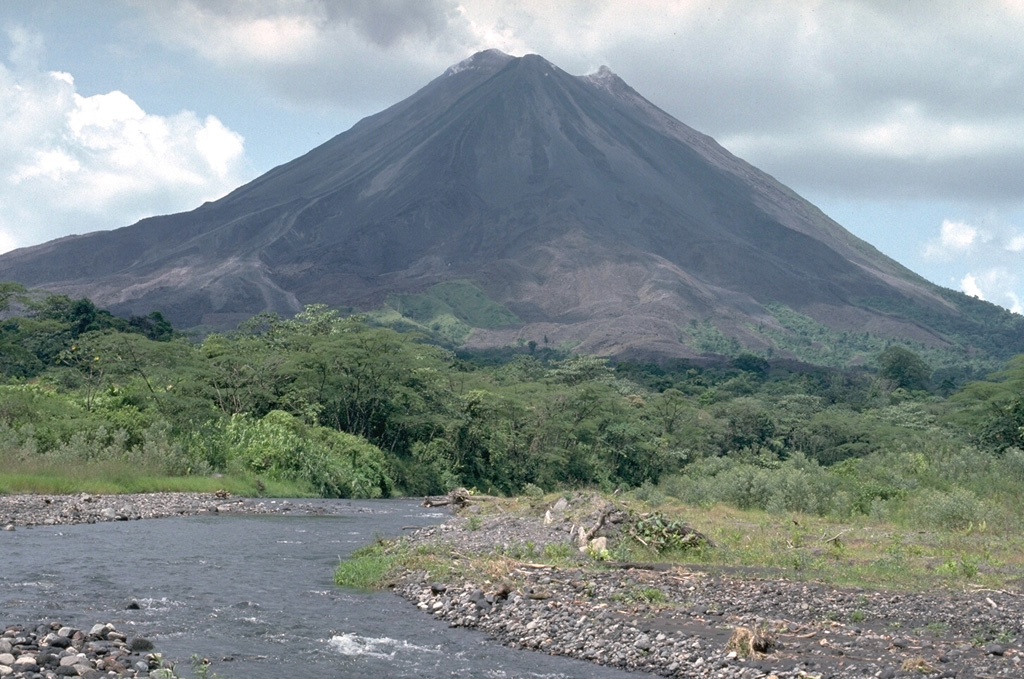 This 1992 view from the SW looks towards Arenal with the new active summit crater just right of the pre-1968 summit, the high point to the left. The 1968-onwards eruption constructed the new cone to the west of the crater from the previous major eruption in about 1525 CE. Lava flows from this eruption cover the unvegetated lower western and southern flanks.    Photo by William Melson, 1992 (Smithsonian Institution)