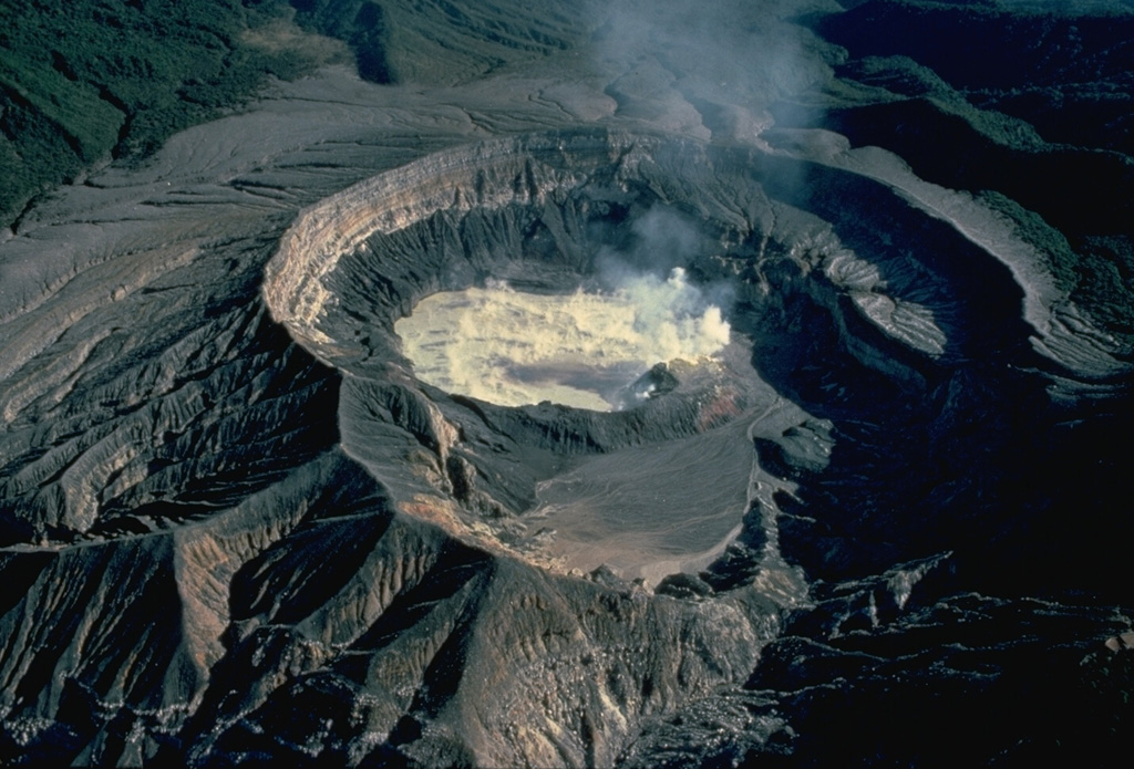 Steam pours from the surface of the hot, acid lake in the active vent of Poás volcano in this 1983 view from the south.  The frequent eruptions from this lake in historical time make Poás one of Costa Rica's most active volcanoes and keep the flanks of the 1-km-wide, 275-m-deep crater vegetation free.  Smaller eruptions produce geyser-like activity within the crater lake.  Occasional larger eruptions completely eject the crater lake. Copyrighted photo by Katia and Maurice Krafft, 1983.