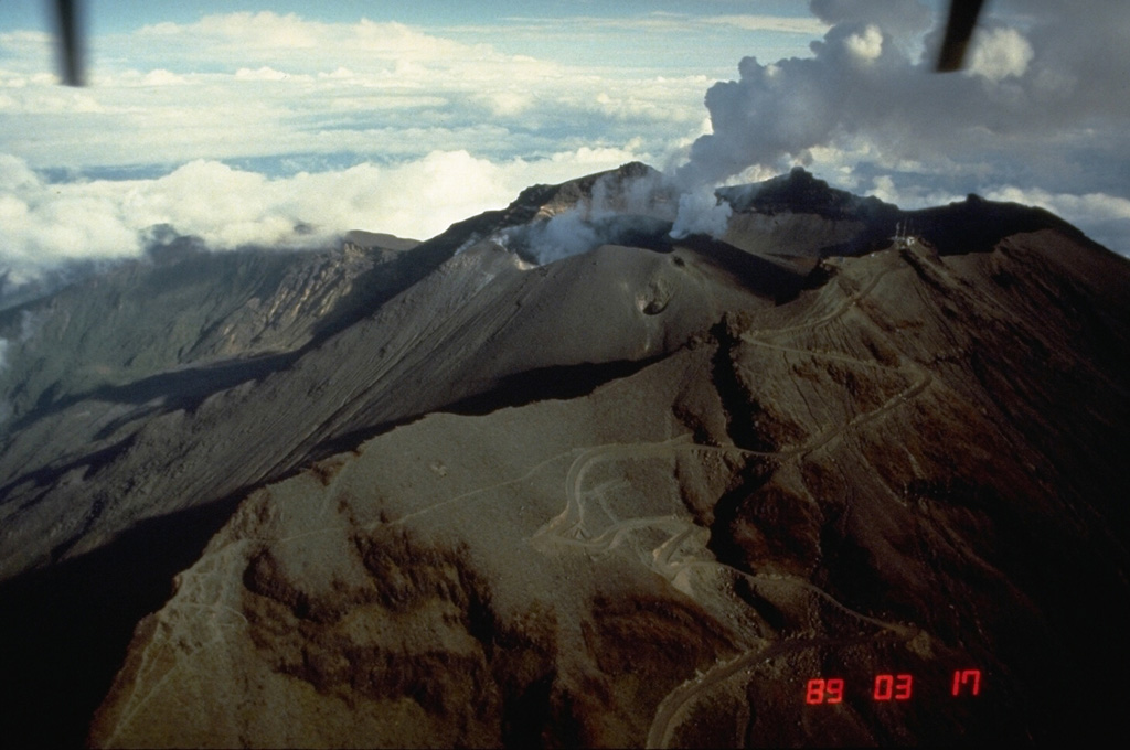 Galeras, a stratovolcano with a large breached caldera located immediately west of the city of Pasto, is one of Colombia's most frequently active volcanoes.  It is seen here from the south on March 17, 1989, with steam clouds pouring from vents on the large central cone near the back headwall of the caldera, whose south rim forms the ridge in the foreground.  Major explosive eruptions since the mid Holocene have produced widespread tephra deposits and pyroclastic flows that swept all but the southern flanks.   Photo by Norm Banks, 1989 (U.S. Geological Survey).