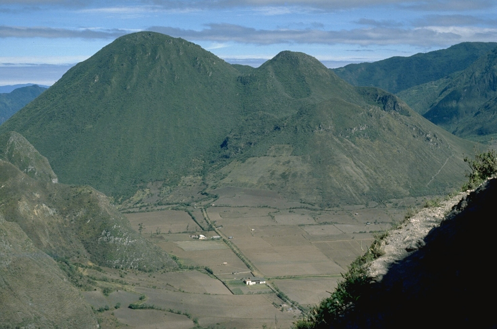 Pululahua is a relatively low-profile, forested volcano immediately north of the equator, 27 km N of Quito. Loma Pondona (left) and the lower Rumiloma (right center) are two of a group of lava domes that partially fill a 3-km-wide caldera. They are seen here from the SE caldera rim. The caldera was formed during the latest dated eruption about 2,400 years ago. Large explosive eruptions were accompanied by pyroclastic flows during the late Pleistocene and Holocene. Photo by Lee Siebert, 1978 (Smithsonian Institution).