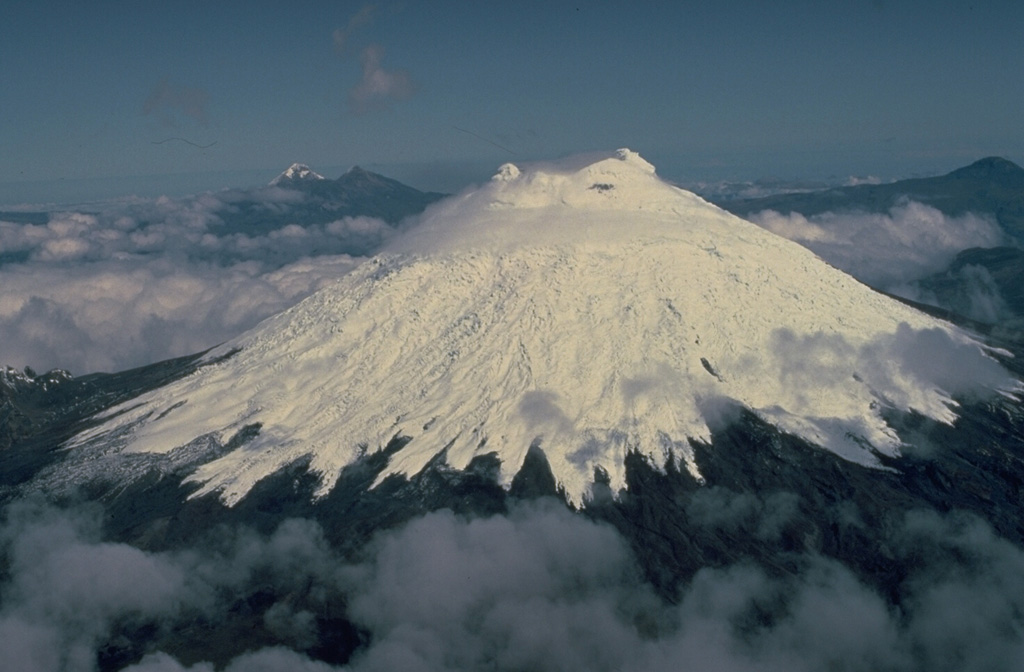 Glacier-clad Cotopaxi volcano is seen here from the SE, with twin-peaked Illiniza volcano in the distance left of the summit.  Cotopaxi is one of Ecuador's most active volcanoes and has erupted more than 50 times since its first historical eruption in the 16th century.  The upper 1400 m of the volcano is ice covered, which has contributed to the production of lahars that have devastated valleys far beyond the volcano. Copyrighted photo by Katia and Maurice Krafft, 1983.