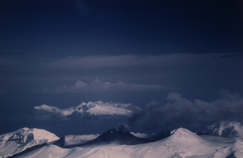 An ash plume rises above the scoria cone in the Akutan summit caldera of on 2 March 1987. This view from the WSW shows Mount Gilbert, a Pleistocene volcano on Akun Island and the peaks of Westdahl, Shishaldin, and Isanotski (left to right) on distant Unimak Island. Photo by Harold Wilson (Peninsula Airways), 1987 (courtesy of John Reeder, Alaska Div. Geology & Geophysical Surveys).