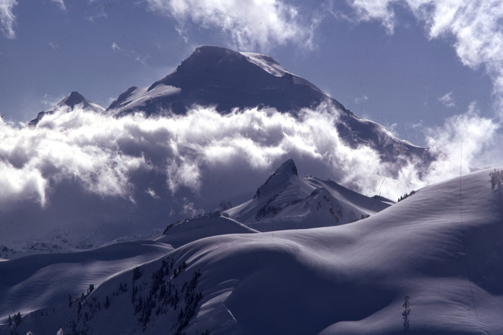 The historically active crater of Mount Baker, seen here from the NE rim of the Pleistocene Kulshan Caldera, lies between the summit and Sherman Peak (left horizon). The snow-mantled slopes in the foreground are located within Kulshan Caldera, which remained undiscovered until the 1990s because associated pyroclastic flow and ashfall deposits outside the caldera had been removed by Pleistocene glaciers. Coleman Pinnacle (center) is a remnant of a post-caldera fissure-fed lava flow. Photo by Lee Siebert, 1974 (Smithsonian Institution).
