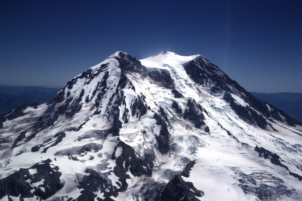 The Tahoma Glacier flows from the summit icecap of Mount Rainier between Liberty Cap (left) and Point Success (right) in this aerial view from the SW in 1969. The current summit was constructed within a scarp left by the collapse of the summit about 5,600 years ago. Slope failure of the summit or upper flanks of the hydrothermally altered volcano has occurred several times during the Holocene, producing massive debris avalanches and mudflows that swept into the Puget lowlands. Photo by Lee Siebert, 1969 (Smithsonian Institution).