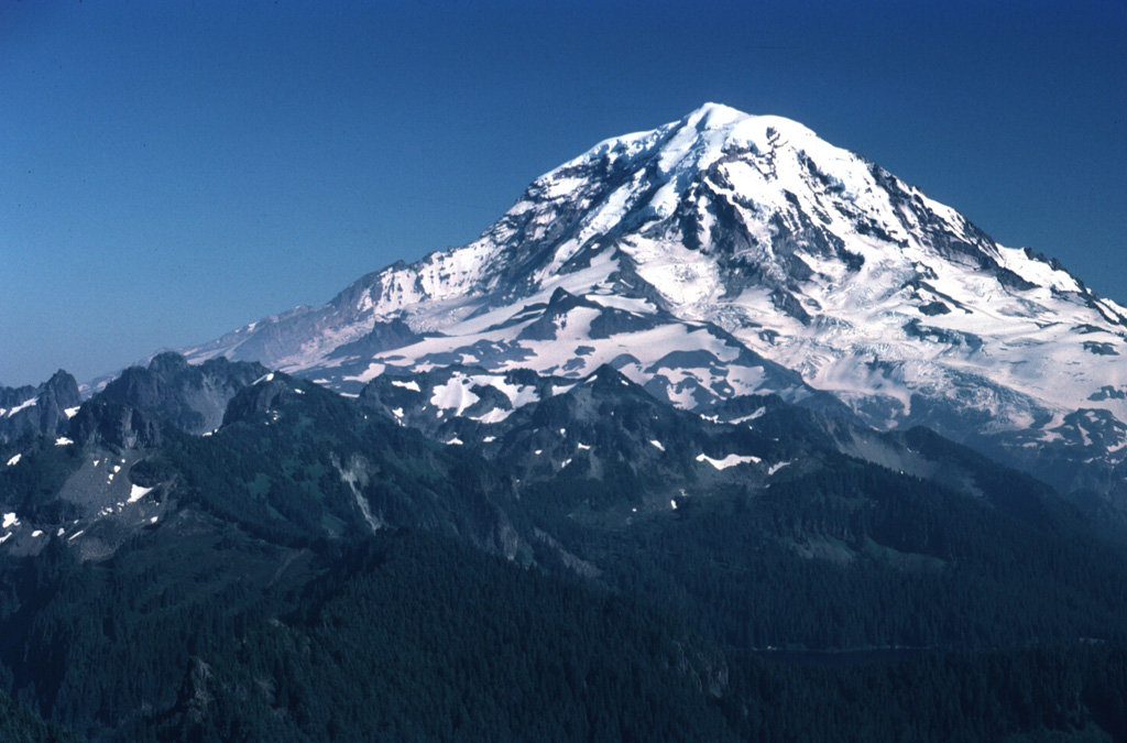 Stratovolcanoes are composed of accumulated layers of lava flows from effusive eruptions and fragmented rock from explosive eruptions. Glacier-clad Mount Rainier, seen here from the NW, is located in the northern Cascade Range. Most eruptions originate from a central conduit, which produces the common conical profile of stratovolcanoes, but flank eruptions also occur. Both isolated stratovolcanoes like Mount Rainier and compound volcanoes formed by overlapping cones are common. Photo by Lee Siebert, 1983 (Smithsonian Institution).