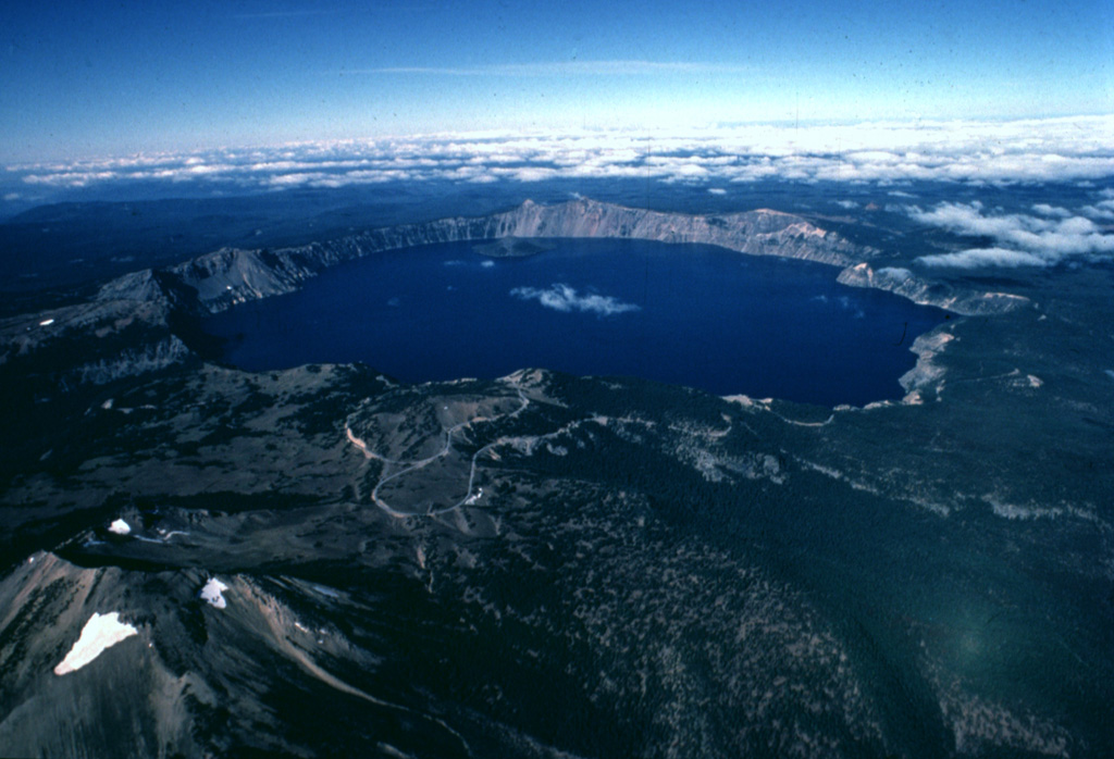 The dramatic 8 x 10 km wide Crater Lake caldera is seen here in an aerial view from the east.  The caldera was formed about 6850 years ago during a major eruption that caused the collapse of ancestral Mount Mazama, a complex of overlapping stratovolcanoes and shield volcanoes.  Mount Scott, a pre-collapse stratovolcano, is the peak with small snowfields at the lower left.  Wizard Island, a post-caldera cinder cone, can be seen near the far side of Crater Lake. Copyrighted photo by Katia and Maurice Krafft, 1984.