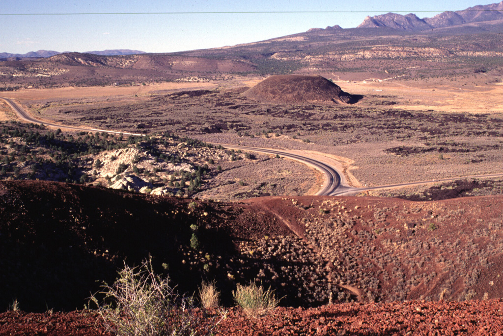 Reddish scoria caps the circular rim of the southernmost of two cinder cones in the lower Diamond Valley of SW Utah.  The northern cone in the middle distance is surrounded by a basaltic lava field.  Light-colored rocks of the Navajo Sandstone at the left lie across a narrow gap from the southern cone; lava flows from the two cones spilled through this gap into Snow Canyon.  The Pine Mountains rising in the distance are in part covered by older lava flows in the St. George basin. Photo by Lee Siebert, 1996 (Smithsonian Institution).