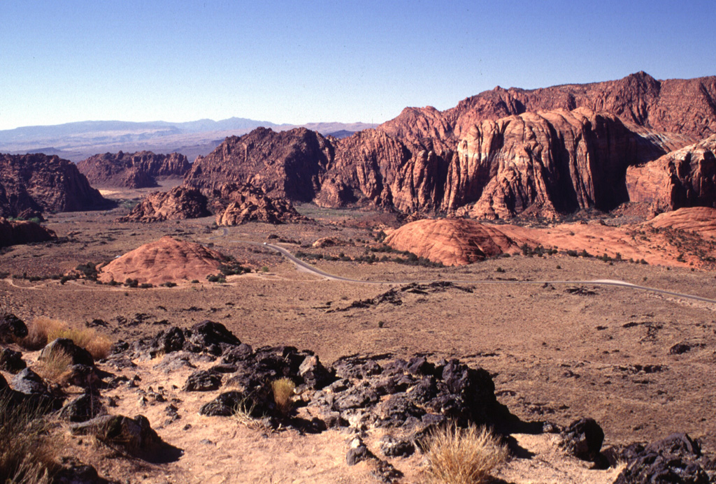 The Santa Clara lava flow fills the valley of Snow Canyon, cut in colorful rocks of the Navajo Sandstone.  The young flow, covered only by scrubby vegetation, split into several lobes and flowed around several peninsulas and islands of the reddish Navajo Sandstone that protrude through the black basalt.  The flow traveled through the narrow gap at the upper left and reached nearly to the Santa Clara River, 16 km from the source of the flow. Photo by Lee Siebert, 1996 (Smithsonian Institution).