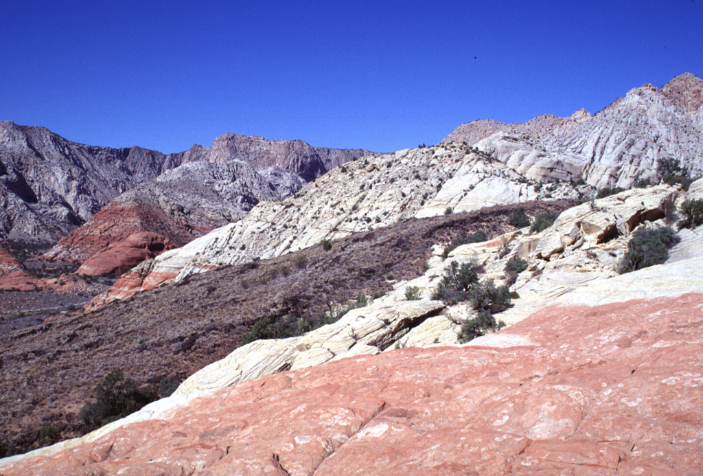 The dark-colored Santa Clara lava flow cutting diagonally across the photo to the left poured over cliffs of the Navajo Sandstone in lava cascades up to 120 m high.  Several lava cascades occurred around sandstone islands in Snow Canyon State Park.  The colorful Navajo cliffs display a transition between underlying reddish oxidized and overlying whitish unoxidized rocks. Photo by Lee Siebert, 1996 (Smithsonian Institution).