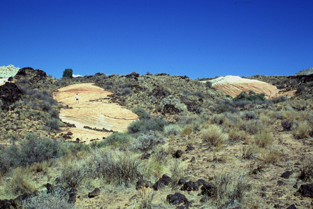 The Santa Clara lava flow in SW Utah split into several lobes and diverted around two islands (kipukas) of colorful cross-bedded Navajo Sandstone.  Note the person standing on the left kipuka for scale.  The largest kipuka of the Santa Clara flow is about 1 km in length. Photo by Lee Siebert, 1996 (Smithsonian Institution).