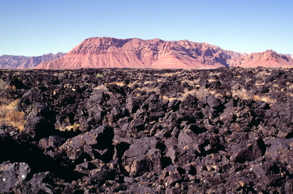 The Santa Clara lava flow exited the narrow Snow Canyon cut in cliff-forming rocks of the Navajo Sandstone in the background.  When the basaltic lava flow reached the flat-lying valley of the Santa Clara River cut in less resistant shales of the Chinle formation, it spread out laterally, reaching a width of more than 1.5 km.  The flow, one of the youngest in Utah, traveled a distance of 16 km from its source in the Diamond Valley.  Housing developments near St. George, Utah now encroach on its distal margins. Photo by Lee Siebert, 1996 (Smithsonian Institution).