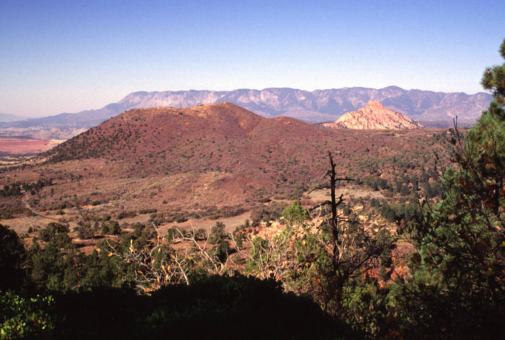 Firepit Knoll (left) is the largest and most well-preserved of a cluster of late-Pleistocene cinder cones on the upper Kolob Plateau in SW Zion National Park.  These cones were erupted through sedimentary rocks of the Navajo Sandstone, which forms the light-colored peak of Red Butte at the right.  The cinder cones erupted basaltic lava flows that traveled down valleys as far as 10-15 km to the south.  The flat-topped summits of the Pine Valley Mountains form the skyline. Photo by Lee Siebert, 1996 (Smithsonian Institution).