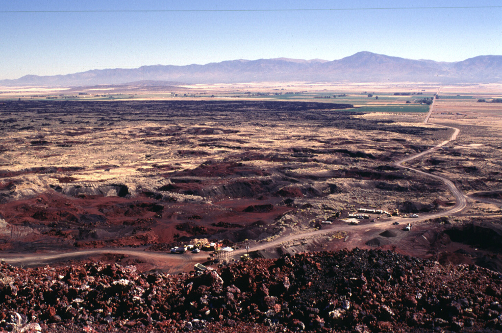The summit of Ice Springs volcano provides a perspective of the extensive lava field erupted from the volcano about 660 years ago.  Ash and scoria from Crescent Crater on the east side of the cone complex was blown primarily to the east by prevailing winds and forms the lighter-colored material that mantles the lava flow in this direction.  The eastern lava flows were erupted early during the Ice Springs eruption.  The road in the foreground provides access to a quarrying operation at the cinder and spatter cone complex.  Photo by Lee Siebert, 1996 (Smithsonian Institution).