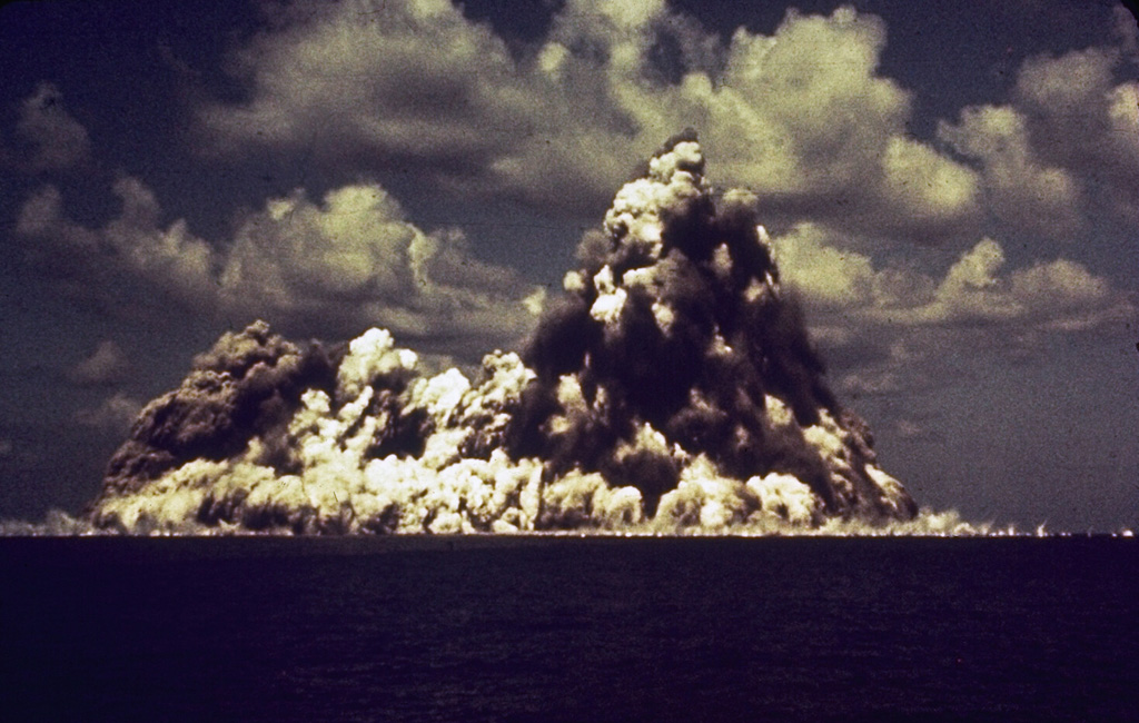 A submarine eruption at Myojinsho lava dome on the rim of Beyonesu Rocks caldera in the central Izu Islands on 23 September 1952. This photo shows a vertically rising plume of ash and steam (cocks’s-tail plume) and a base surge traveling across the sea surface. This photo was taken 8 seconds after the explosion first breached the surface. The eruption began on 16 September when a lava dome rose towards the sea surface. Explosive activity then progressively destroyed the dome. This cycle occurred three times until October 1953. Photo courtesy of Ryohei Morimoto (University of Tokyo).