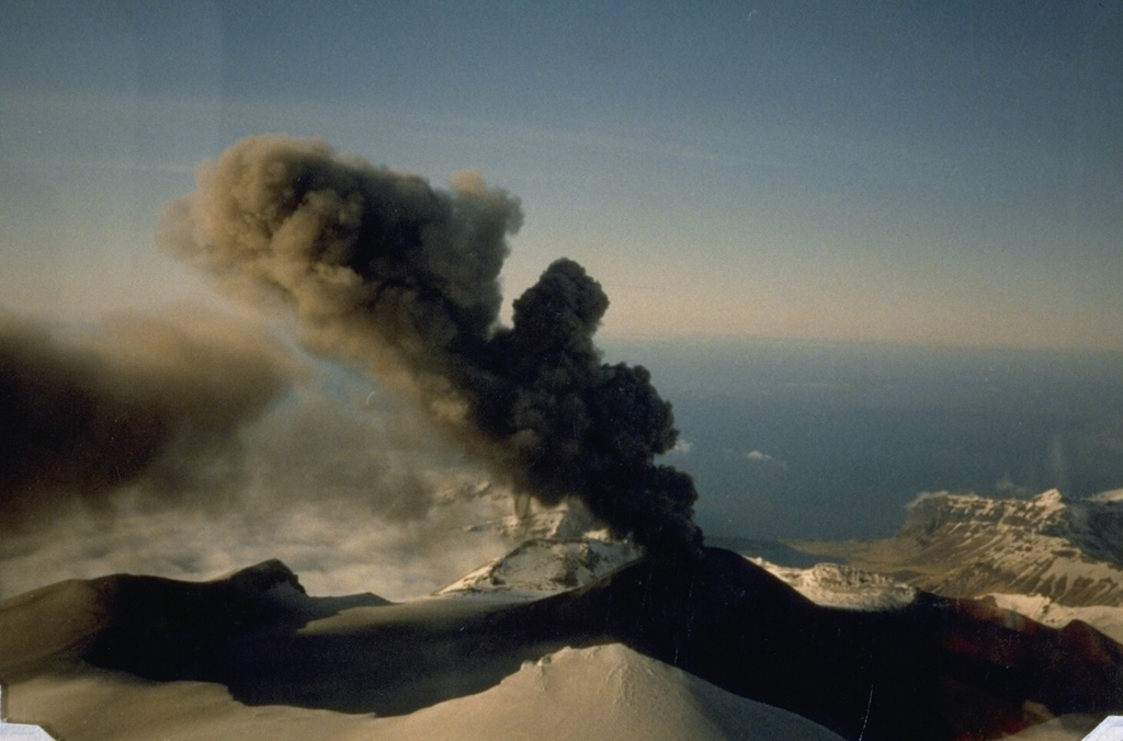 An ash plume rises about 1 km and drifts to the NW above the Akutan caldera scoria cone on 2 June 1988. Dark ash deposits on the flanks extend across the caldera floor and western rim. The snow-covered ridge in the foreground is the SW caldera rim. Intermittent eruptions such as these are typical of Akutan. The 1988 eruption lasted from 26 March to 20 July. Photo by Tom Madsen (Aleutian Air), 1988 (courtesy of John Reeder, Alaska Div. Geology & Geophysical Surveys).