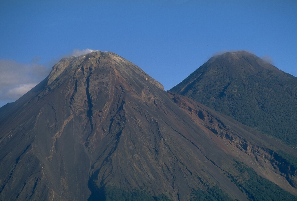 Fuego and Acatenango are seen here from the SE and are result of eruptions along a N-S line that moved progressively towards the south. The gully on the righthand side of Fuego is the rim of a scarp produced by the collapse of ancestral Meseta volcano, which preceded the growth of the modern Fuego edifice. Copyrighted photo by Stephen O'Meara, 1994.