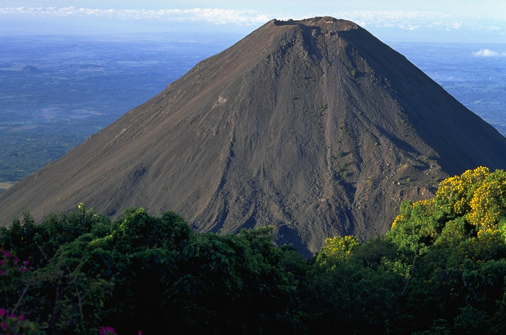 Volcán de Izalco is seen here from the north, on the southern flank of Santa Ana. It began erupting in 1770 CE and frequent Strombolian eruptions produced a steep-sided, 650-m-high stratovolcano over a 200-year period. Izalco was one of the most frequently active volcanoes in Central America, producing ejecta and lava flows from both summit and flank vents. Copyrighted photo by Stephen O'Meara.