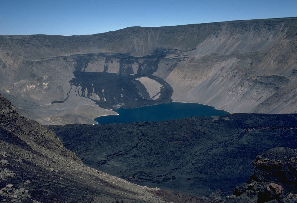 The dark lava flows at the far NW end of Fernandina caldera were emplaced in August 1978.  An eruption beginning on August 8 produced an eruption cloud 4.5-6 km high.  This was followed by the emplacement of a large lava flow from a 1-km-long fissure system on the NW caldera bench that flowed into the caldera lake.  Lava fountaining was observed during visits to the caldera rim, on August 10-13 and 16-19.  The eruption had ended by the 26th.  Fresh lava flows in the foreground mantle the surface of the prominent SE caldera bench. Photo by Lee Siebert, 1978 (Smithsonian Institution).
