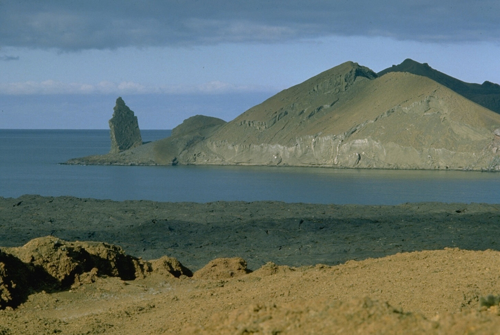 A spectacular suite of volcanic features flanks Sullivan Bay, at the eastern end of Santiago Island.  This view looks NE across the bay to Bartolomé Island.  The sharp pinnacle at the left, a remnant of an eroded tuff cone, is one of the many scenic highlights of the Galápagos Islands.  Bartolomé Island contains eroded tuff cones, a dissected solidified lava lake, and a youthful lava plateau.  The flat coastline of Santiago Island in the foreground is formed from a vast field of youthful pahoehoe lava flows that wraps around the SE corner of the island. Photo by Lee Siebert, 1978 (Smithsonian Institution).