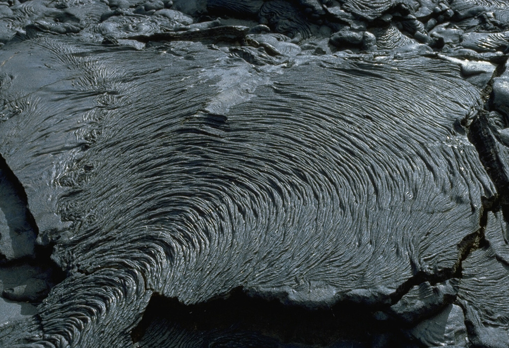 The ropy texture of pahoehoe lava flows is produced when the thin solidifying surface of the flow is pushed by the advancing, still-molten interior. This pleated pahoehoe lobe, on a lava flow at Santiago shield volcano in the Galápagos Islands, advanced slowly from the bottom left to the top right. Pahoehoe lavas are the least viscous of common lava types, and thus form diverse surface structures. Photo by Lee Siebert, 1978 (Smithsonian Institution).