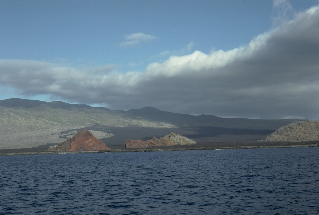 The elongated shield volcano of Santiago Island is dotted with Holocene pyroclastic cones.  Fresh lava flows that blanket the flanks of the volcano originated from these cones.  The 920-m-high summit ridge, lined with NW-trending cinder and spatter cones, is seen here from James Bay on the west side of the island.  The James Bay lava flows (center) reached the coast along a broad front.  They were dated by fragments of marmalade pots left by buccaneers in 1684 that were subsequently embedded in the lava flows observed by Charles Darwin in 1835.  Photo by Lee Siebert, 1978 (Smithsonian Institution).