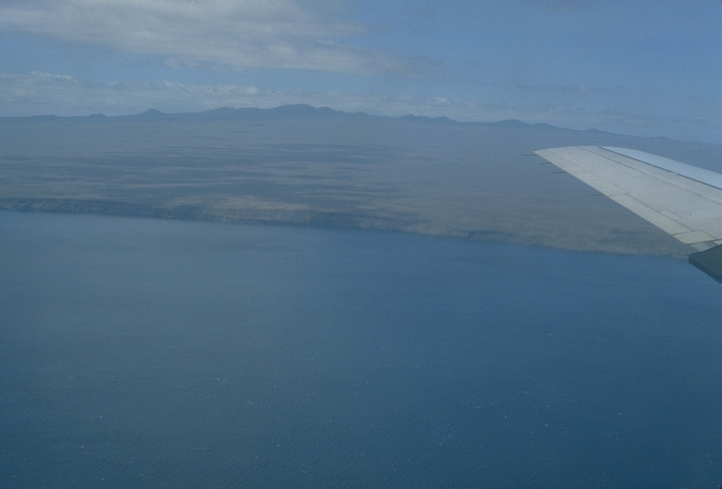 The highlands of the broad Santa Cruz shield volcano, seen here from the NE, are capped by youthful cinder cones with well-preserved craters.  The scoria cones are grouped in an E-W belt parallel to recent fault scarps that border Academy Bay and largely bury a shallow summit caldera.  Older uplifted submarine lava flows are found on the NE part of the island and at the fault-delimited offshore island of Baltra.  No historical eruptions are known from Santa Cruz, the 2nd-most populated island of the Galápagos archipelago. Photo by Lee Siebert, 1978 (Smithsonian Institution).