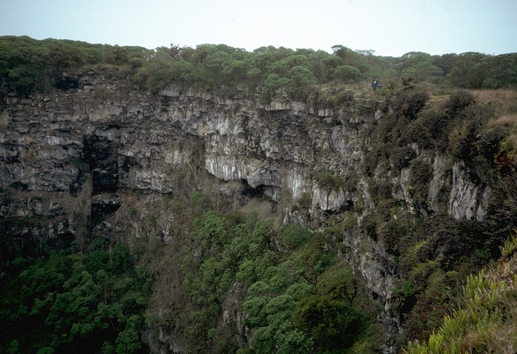 Lava flows line the steep walls of a pit crater on Santa Cruz island.  Note the person standing on the rim at the right for scale.  Pit craters are formed by collapse following the withdrawal of magma along a rift zone.  They differ from other craters in that their rims lack a mantle of explosive debris.  In some cases, vertical-walled pit craters can be hundreds of meters deep. Photo by Lee Siebert, 1978 (Smithsonian Institution).