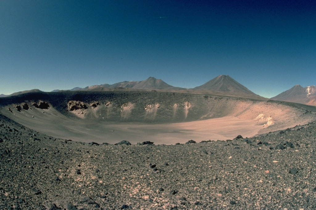 The solitary Holocene Cerro Overo maar, seen here from the south, was erupted through basement ignimbrites of Pliocene age, which form the light-colored rocks in the crater wall.  A thin blanket of dark ejecta from the eruption that created the 600-m-wide, 80-m-deep maar caps its rim.  Pleistocene volcanoes of the central Chilean Andes form the horizon. Copyrighted photo by Katia and Maurice Krafft, 1983.
