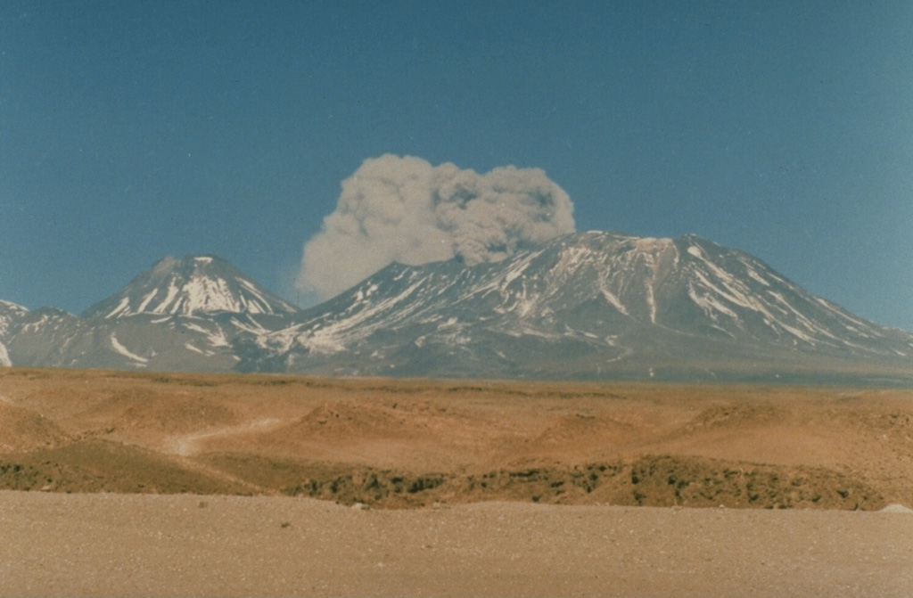 The onset of a brief explosive eruption from Láscar volcano is seen here at about 1430 hrs on September 14, 1986, from Toconao, 33 km NW.  The September 14 eruption produced ash clouds that rose a few hundred m above the vent for about a half hour.  Similar activity on the 15th was followed by a brief, but powerful explosion on the 16th that deposited ash in Salta, Argentina, 350 km to the SE.  Conical Volcán Aguas Calientes rises to the left of the plume. Photo by Paul King, MINSAL Corporation, 1986 (courtesy of Peter Francis, Open University).