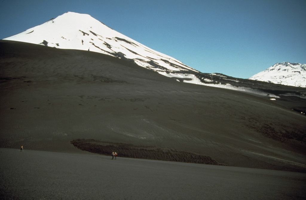 Lonquimay (left) is a small, flat-topped, symmetrical stratovolcano of late-Pleistocene to dominantly Holocene age located immediately SE of the largely Pleistocene Tolguaca volcano (extreme right).  The Cordón Fissural Oriental fissure zone extends 10 km NE of Lonquimay and has produced a series of  vents and cinder cones that have been the source of voluminous lava flows in historical time.  Major lava flows erupted during 1887-90 and 1988-90 traveled up to 10 km from their NE-flank vents.  Photo by Norm Banks, 1990 (U.S. Geological Survey).