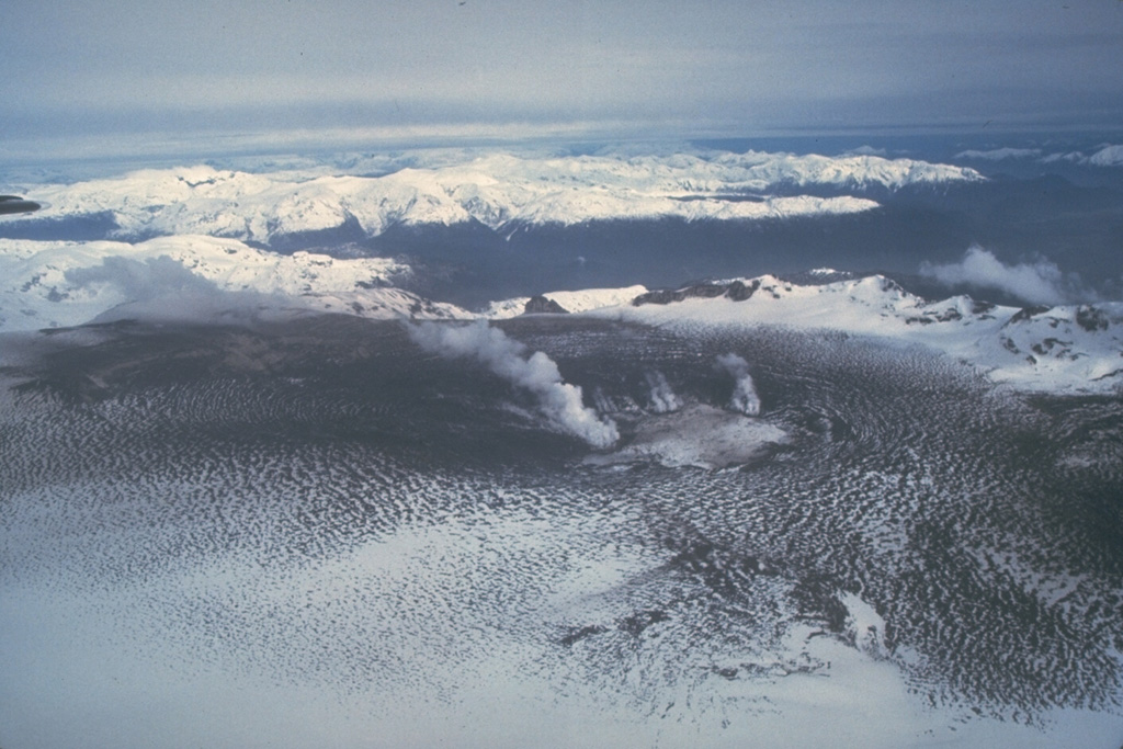 Ash from the 1991 eruption darkens the glacier filling the caldera of Hudson volcano.  This August 23 view from the NE shows steam clouds rising from the margins of a 800-m-wide subglacial crater in the SW part of the caldera that was formed during violent explosive eruptions August 12-15.  Ashfall from the eruption spread eastward across Argentina, damaging airport facilities, houses, and agricultural land.  The eruption caused major mortality of grazing animals, whose food and water were contaminated by fluorine-rich ashfall. Photo by Norm Banks, 1991 (U.S. Geological Survey).