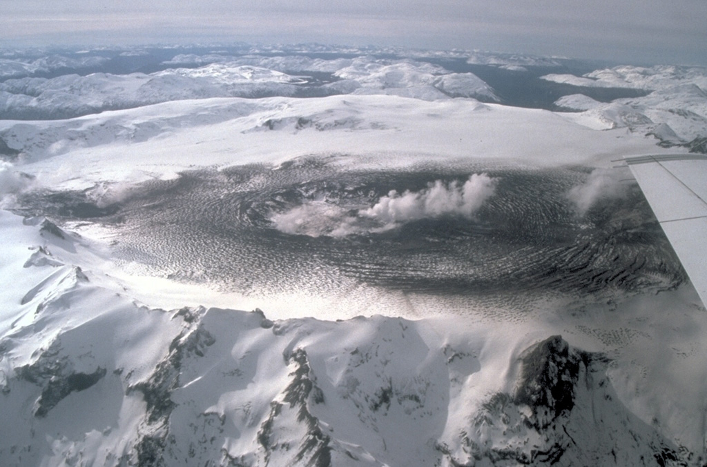 The ice-filled 10-km-wide caldera of the remote Cerro Hudson volcano was not recognized until its first 20th-century eruption in 1971.  The massive, 1905-m-high Cerro Hudson, seen here from the west on August 23 during its 1991 eruption, covers an area of 300 sq km.  The caldera is drained through a breach on its NW rim (upper left), which has been the source of mudflows down the Rio de Los Huemules.  The 1991 eruption was Chile's second largest of the 20th century, and formed a new 800-m-wide crater in the SW part of the caldera.   Photo by Norm Banks, 1991 (U.S. Geological Survey).