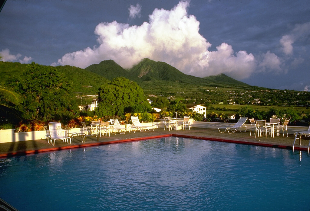 During more pleasant days, Soufrière Hills volcano rises above the outskirts of Plymouth (lower right), the capital city of Montserrat, known as the Emerald Isle.  Two decades after the date of this 1977 photo, pyroclastic flows from a growing lava dome swept through the center of the evacuated city of Plymouth. Photo by Richard Fiske, 1977 (Smithsonian Institution).