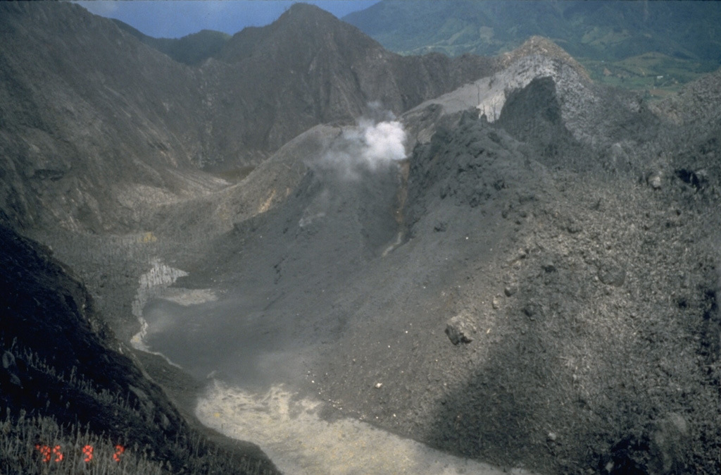 The south moat of English's Crater is seen here from the east on September 2, 1995.  A blanket of ash covers Castle Peak lava dome (right) and the walls of English's Crater.  Steam rises from a vent on the SW side of the lava dome that formed on July 28.  A new lava dome began growing in November 1995.  It eventually grew above the crater rim and filled in most of the moat of English's crater.  Pyroclastic flows initially swept into the sea through the breached eastern end of English's Crater, but later overtopped its rim and flowed down the outer flanks. Photo by Cynthia Gardner, 1995 (U.S. Geological Survey).