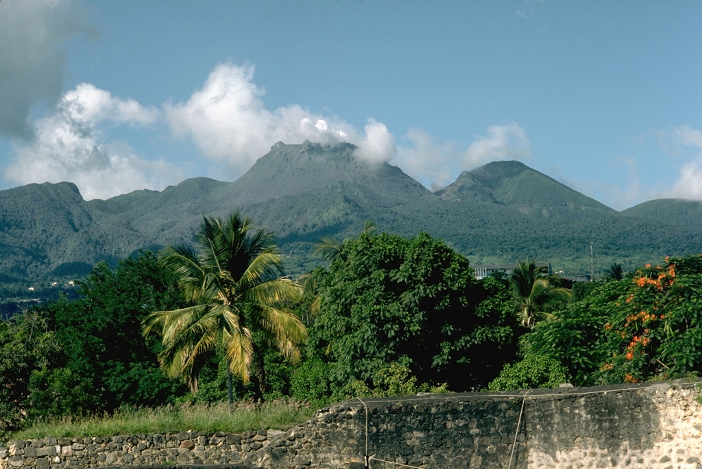 Steam rises from a 500-year-old lava dome that forms the summit of La Soufrière volcano on Guadeloupe.  It was constructed within the 1-km-wide, horseshoe-shaped Amic Crater, whose northern rim is the flat-topped ridge at the extreme left.   Amic Crater was the source of a major explosion and debris avalanche about 3100 years ago during an eruption that resembled the 1980 Mount St. Helens eruption.  The peak to its right is l'Echelle, a young cinder cone.  This September 1976 view is from the west. Photo by Richard Fiske, 1976 (Smithsonian Institution).