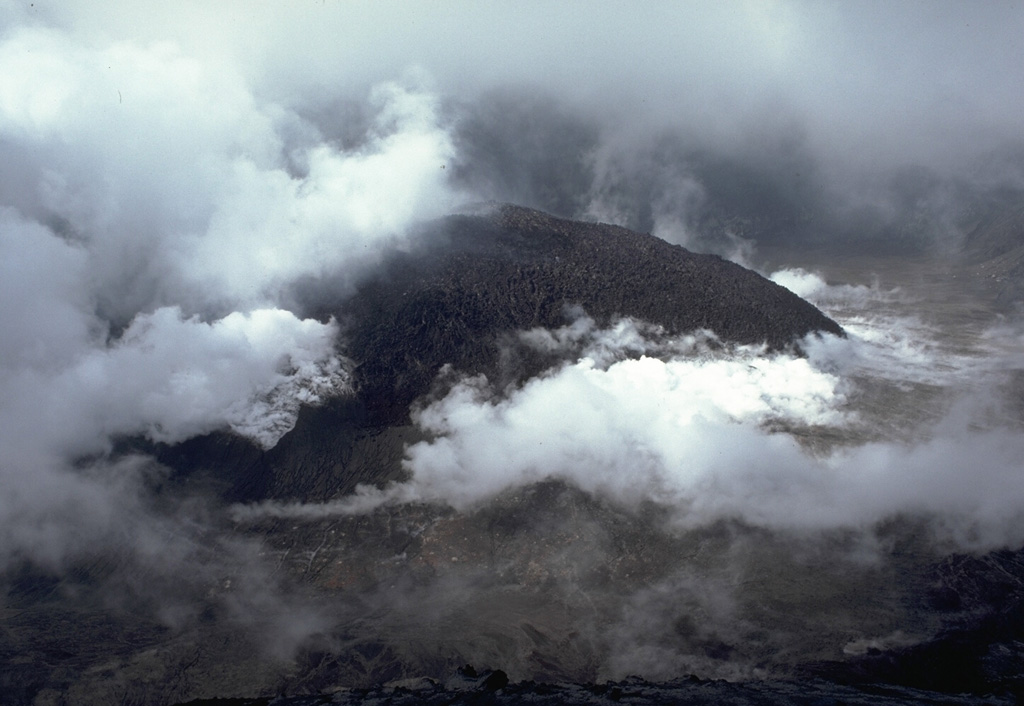 A steaming lava dome fills the floor of the summit crater of Soufrière volcano in June 1979.  Both steam clouds from the degassing lava dome and atmospheric clouds are present in this photo.  The dome began growing on May 3, after the cessation of explosive eruptions on April 25, and reached a height of 110 m by June 18.  By the end of the eruption in October, the dome was 130-m high.   Photo by Richard Fiske, 1979 (Smithsonian Institution).