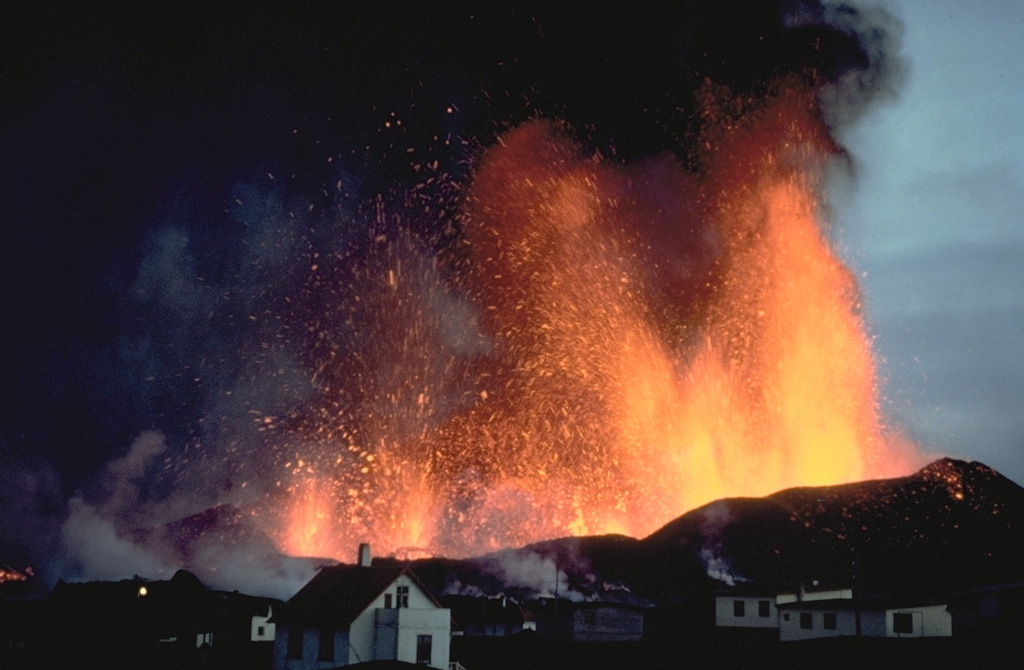 Lava fountains tower above the town of Vestmannaeyjar in March 1973.  The eruption began on January 23, 1973, with explosive ejections from a NNE-SSW-trending, partly submarine fissure cutting Helgafell cone.  Lava fountains built the Eldfell cinder cone and fed lava flows that reached the sea.  Tephra buried more than 100 houses, and lava flows inundated parts of the town and threatened its harbor.  The effects of vigorous efforts to slow the progress of the lava by spraying it with cold seawater are debated, but the flow stabilized at a point that improved the natural harbor.  Copyrighted photo by Katia and Maurice Krafft, 1973.