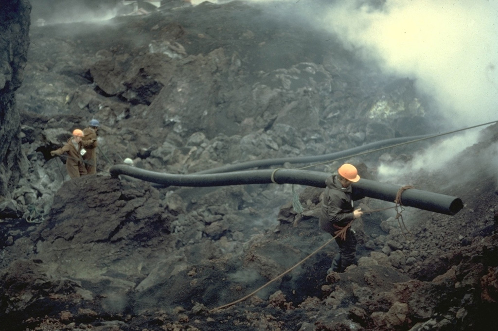 Workers carry pipes used during a massive attempt to cool the advancing lava flow produced by the 1973 eruption of Heimaey volcano.  Water was pumped beginning on February 6, initially using a single large pump, and then larger pumps flown in from Iceland and the United States.  On March 1, a pumping vessel arrived that sprayed water from the harbor.  Cooling locally produced solidified barriers that diverted and stopped the lava flow.  The lava flow created new land when it entered the sea and eventually improved the island's only harbor. Copyrighted photo by Katia and Maurice Krafft, 1973.