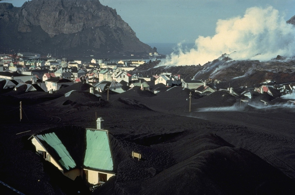 A steaming lava flow advances through the town of Vestmannaeyjar in April 1973.  Houses in the foreground are partly buried by thick ashfall deposits from the Eldfell cinder cone, which began erupting on January 23.  The lava flow advanced into the town's harbor, which lies between the flow and the cliff at the upper left.  The flow covered a 300-m-wide, 700-m-long area in the NE part of town, burying 200 houses and buildings during a two-week period of extrusion in late March and early April. Copyrighted photo by Katia and Maurice Krafft, 1973.