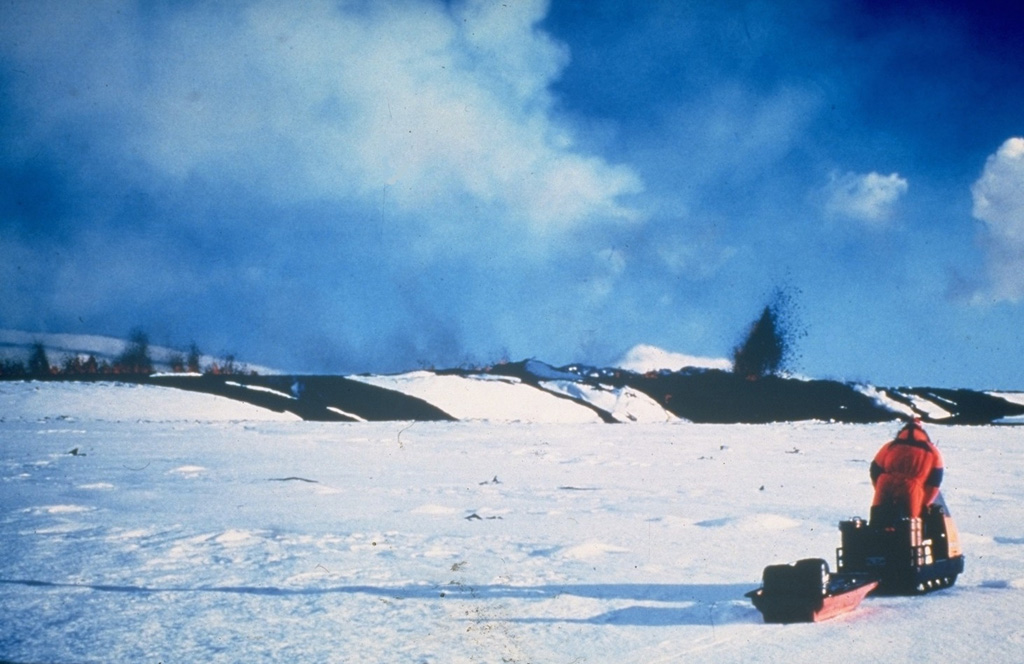 Two scientists of the Nordic Volcanological Institute doing monitoring fieldwork on 16 March 1980 were able to document the onset of activity when deflation started and an eruption began. Small dark lava fountains can be seen in a line on the left, indicating the location of a fissure, with a single larger fountain on the right which may represent a vent on the end of the fissure. Dark lava flows can be seen on the snow. A few minutes after this photo was taken a lava flow rapidly advanced in their direction, prompting them to retreat on their snowmobiles. Photo courtesy of Gudmundar Sigvaldason, 1980 (Nordic Volcanological Institute, Reykjavík).