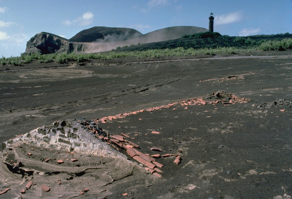 A submarine eruption that began off the western coast of Fayal Island in 1957 produced heavy ashfall that buried houses, such as the one in the foreground.  The new submarine vent built a pyroclastic cone (upper left) that was eventually connected to the mainland.  Extensive ashfall partially buried the Capelinhos lighthouse (upper right), which had been located on the western shoreline of Fayal Island. Copyrighted photo by Katia and Maurice Krafft, 1980.