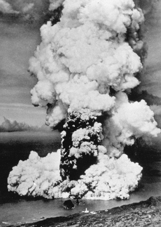 A column of ash and steam rises above a new submarine vent off the western coast of Fayal Island in early October 1957. Horizontally traveling base surges form a ring at the base of the eruption column. Explosive eruptions began on September 27 and formed a small island that eventually was joined to the main island, partially burying the Capelinhos lighthouse, which is visible on the center shoreline (bottom). Lava effusion was continuous for the last five months of the 13-month eruption. Photo by U.S. Air Force, 1957 (published in Green and Short, 1971).