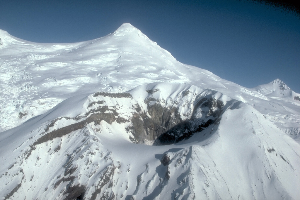 Crater Peak, a flank vent of Mount Spurr, is seen from the south with the snow-and-ice-covered summit lava dome complex in the background. Both the summit lava dome and Crater Peak were constructed within a 5-6 km wide scar formed by an earlier collapse. Crater Peak has been the source of about 40 Holocene tephra layers identified in the Cook Inlet basin. Photo by Game McGimsey, 1991 (Alaska Volcano Observatory, U.S. Geological Survey).
