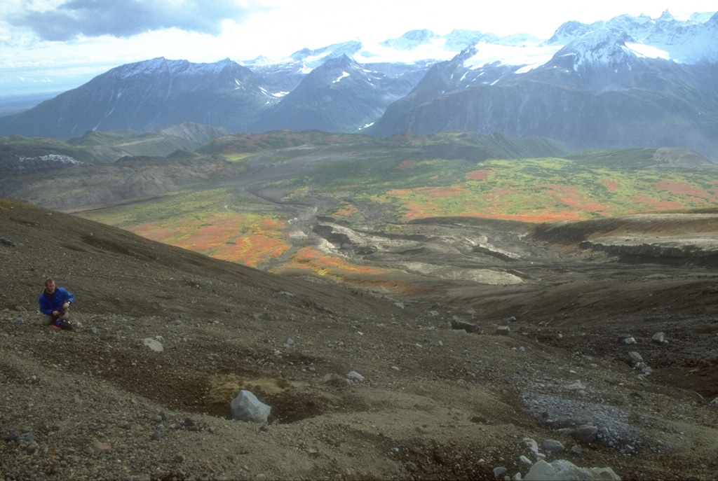 A volcanologist from the Alaska Volcano Observatory inspects an impact crater (left-foreground) formed by a dense block ejected during the 18 August 1992 eruption of the Crater Peak vent on Mount Spurr. Pyroclastic flow and lahar deposits from that eruption form the darker areas descending SE-flank valleys. This 1993 view looking away from Spurr shows glacier-covered plutonic and sedimentary rocks of the Alaska Range in the background. Photo by Christina Neal, 1992 (Alaska Volcano Observatory, U.S. Geological Survey).